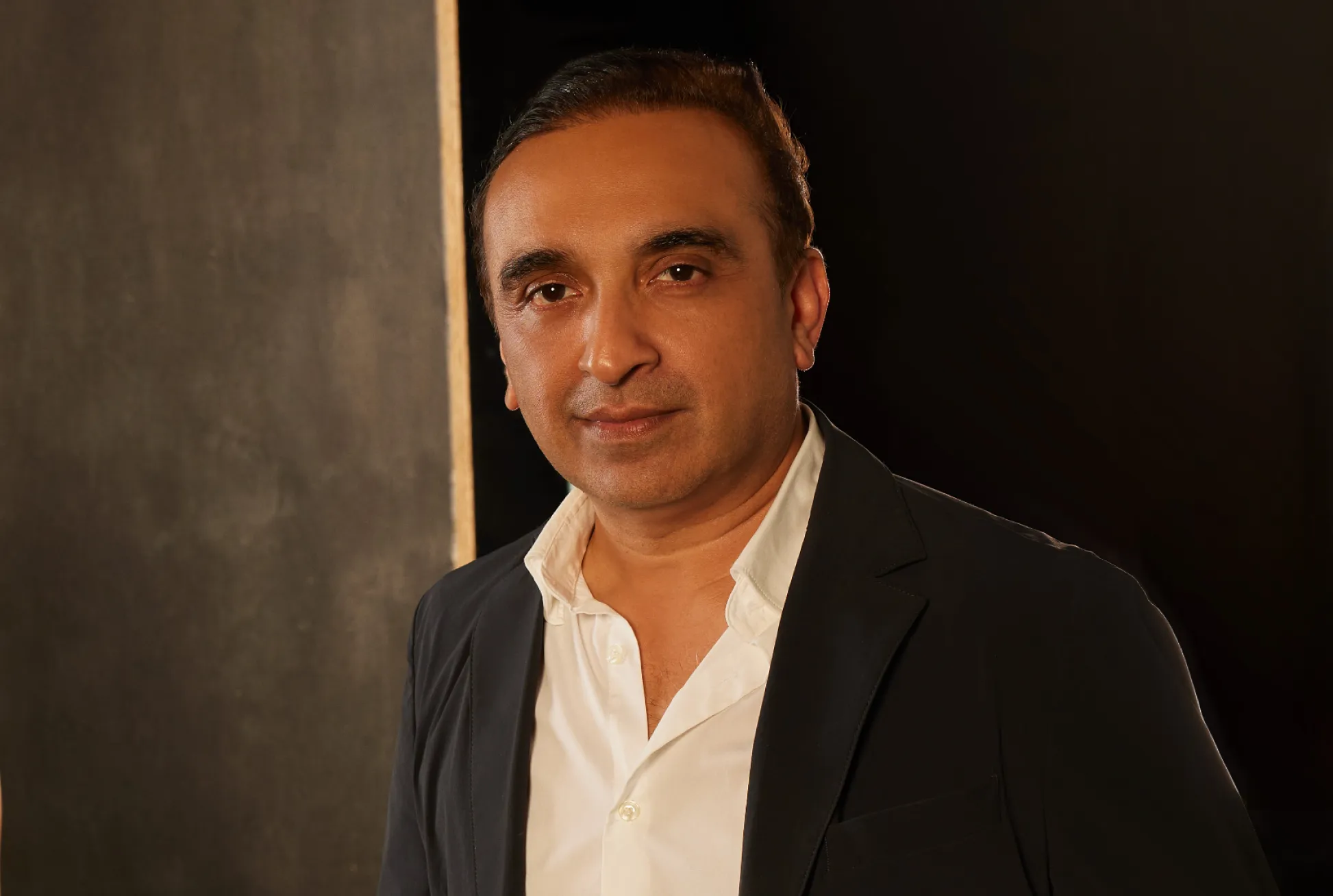 Man in a dark blazer and open white shirt against a dark textured wall