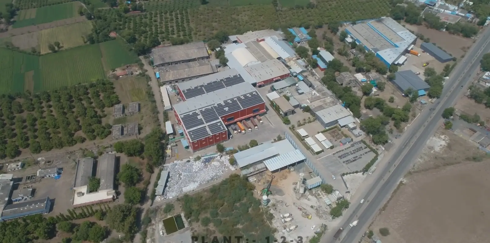Aerial view of a manufacturing plant with rooftop solar panels beside farm fields and a highway