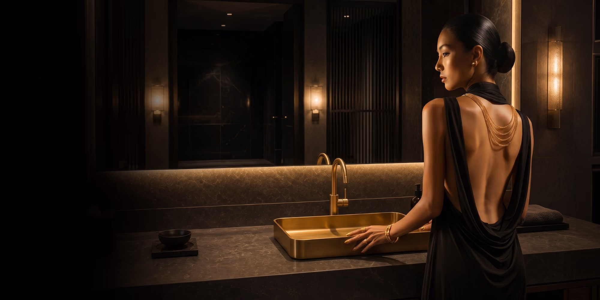 Woman in a black gown at a gold countertop basin with gold tap in a dimly lit bathroom