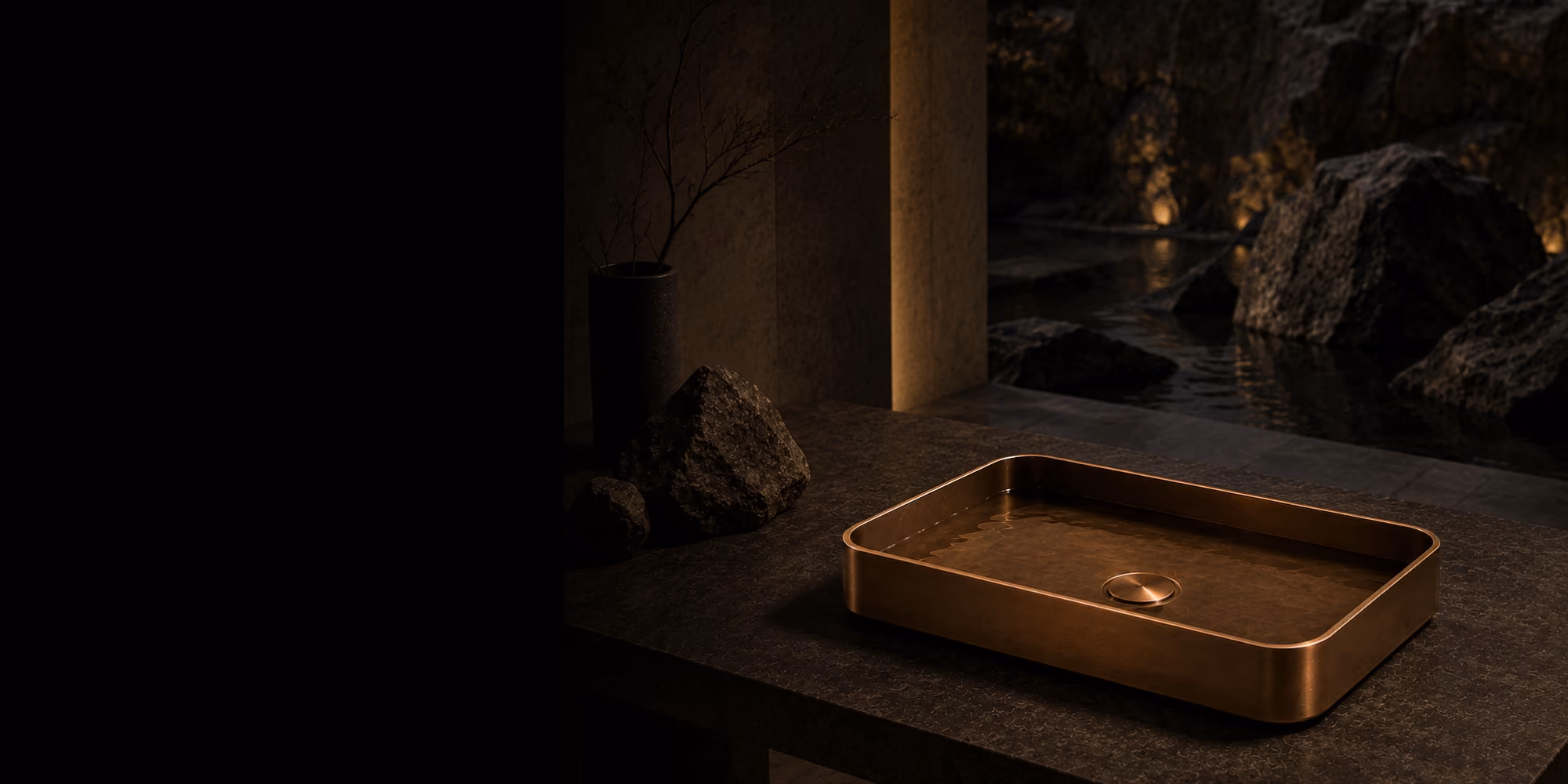 Inox bronze rectangular countertop basin on a dark stone ledge lit against a rocky pool