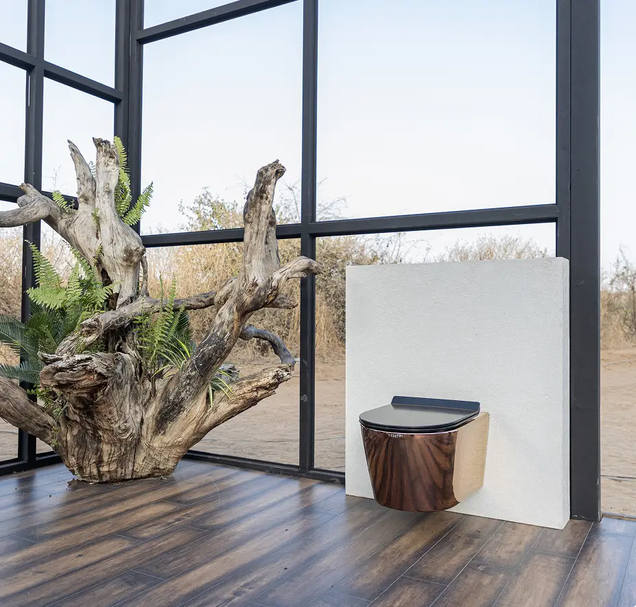 Copper wall-hung toilet on a white panel beside driftwood and ferns in a glass-walled room