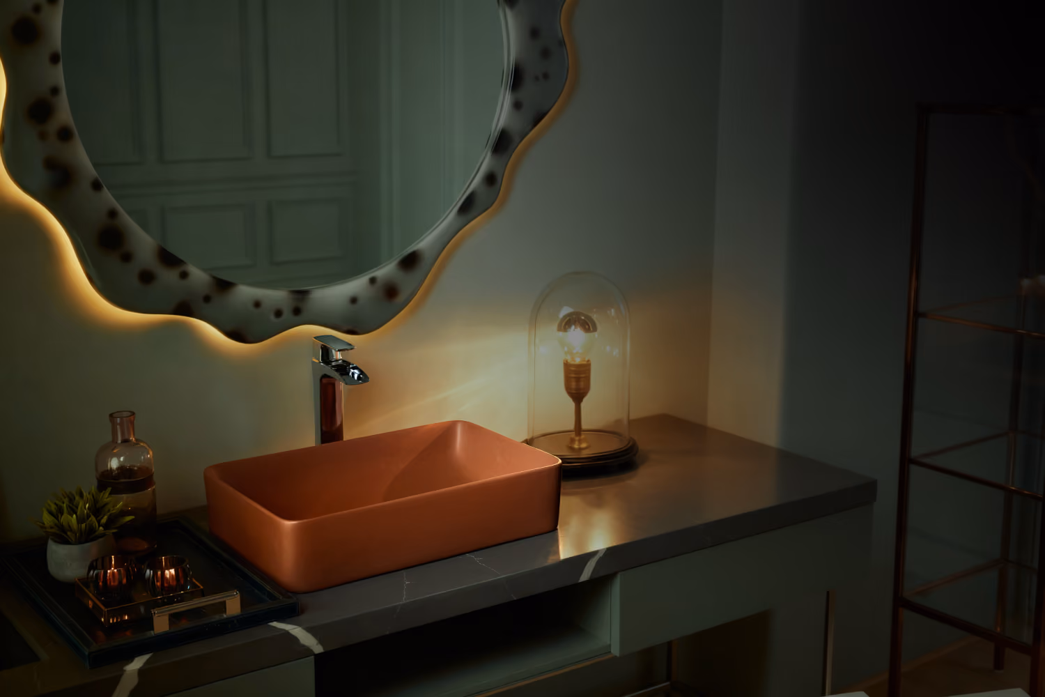Dim green bathroom with terracotta vessel basin, chrome tap, backlit wavy mirror and table lamp