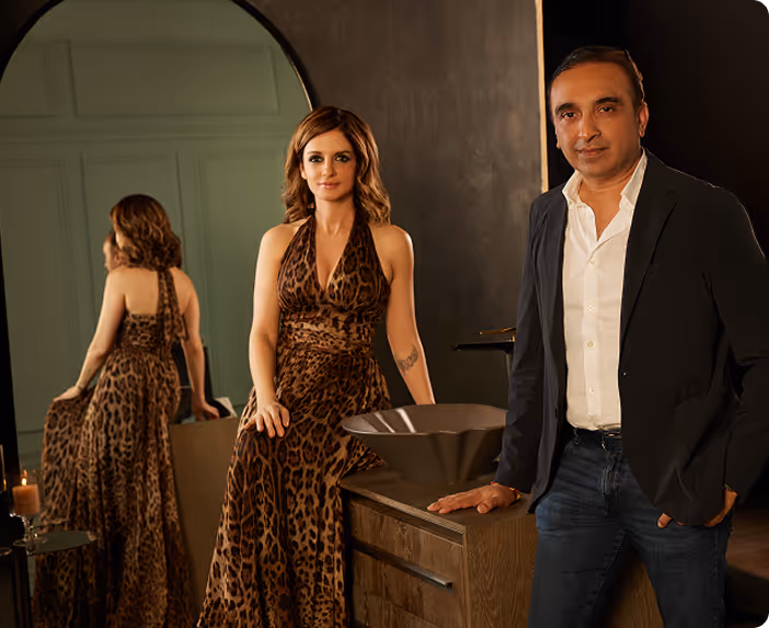 Man and woman posed beside a wooden vanity and arched mirror in a dark bathroom setting
